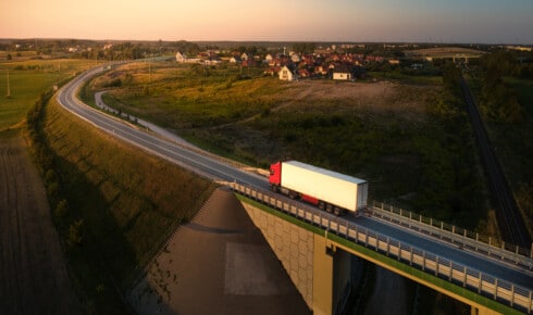 Hvad er HVO100? Luftfoto af en lastbil, der k&oslash;rer ad en landevej ved solnedgang, med en landsby og en bro i et nordisk landskab &ndash; et symbol p&aring; en fl&aring;de, der k&oslash;rer p&aring; fossilfri diesel
