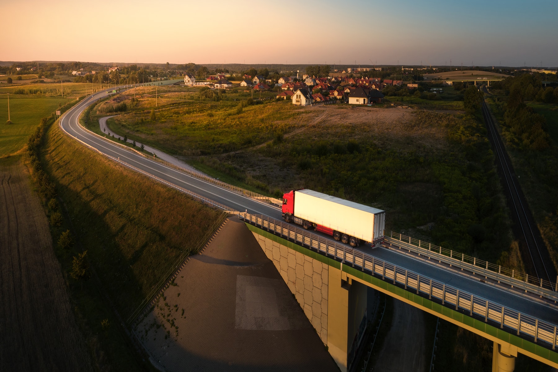 Hvad er HVO100? Luftfoto af en lastbil, der kører ad en landevej ved solnedgang, med en landsby og en bro i et nordisk landskab – et symbol på en flåde, der kører på fossilfri diesel