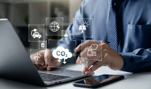 Hvad er Scope 1 2 og 3 emissioner: en fagperson arbejder på en bærbar computer, omgivet af digitale ikoner for CO2-udledning, en fabrik, vedvarende energi og en elbil – et billede på den digitale sporing bag virksomheders emissionsrapportering